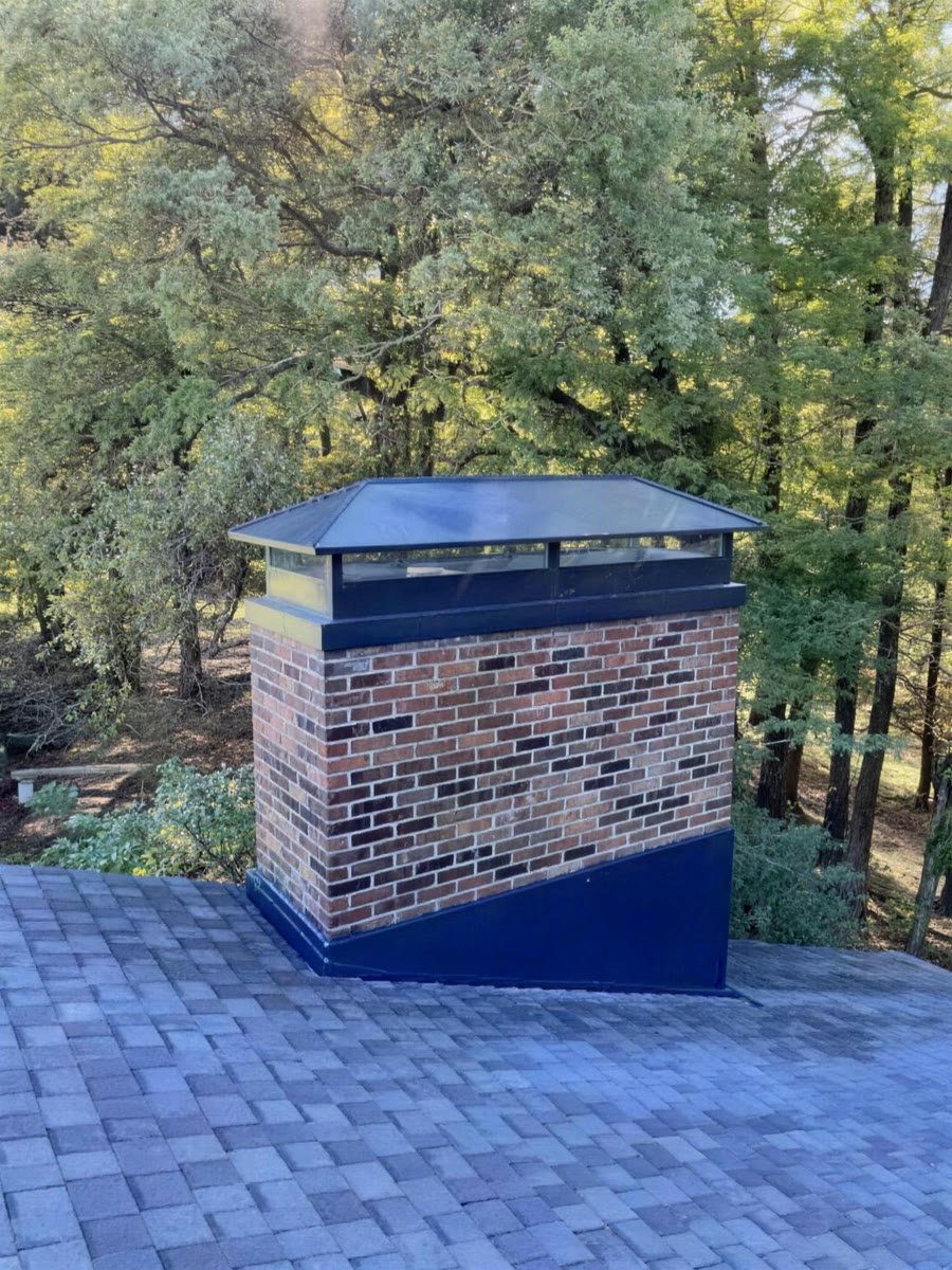 Brick chimney with a custom black metal top and shroud, autumn trees behind